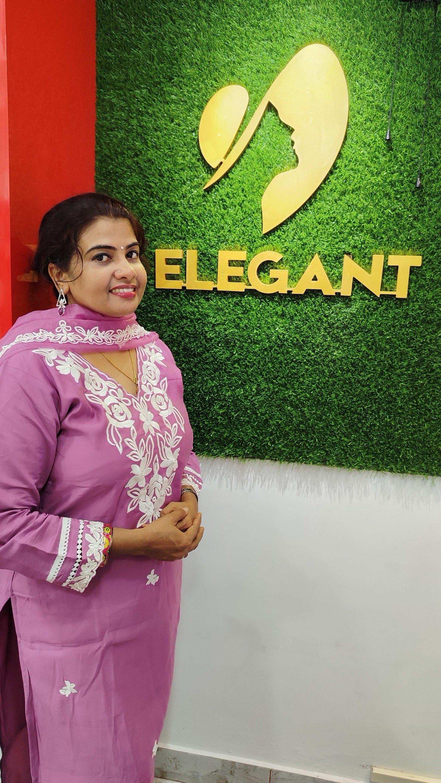 Woman wearing a silk churidar set in pink with white embroidery standing by a green wall with Elegant logo