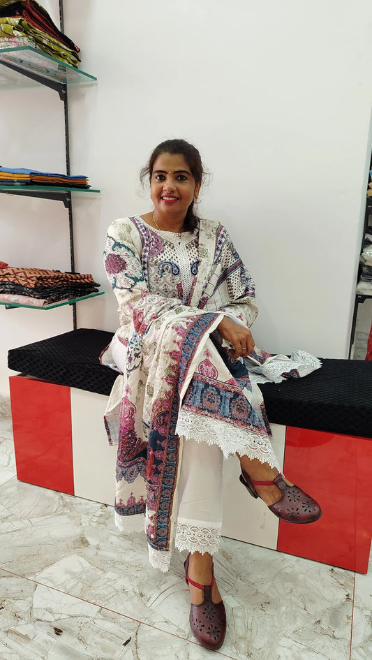 Woman wearing a white Kora Maslin churidar set with multicolored floral and geometric prints sitting on a bench in a fabric store.