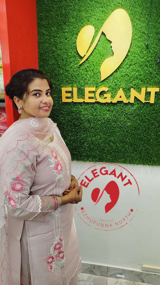 Woman in light pink floral embroidered silk churidar set posing indoors near elegant store logo sign