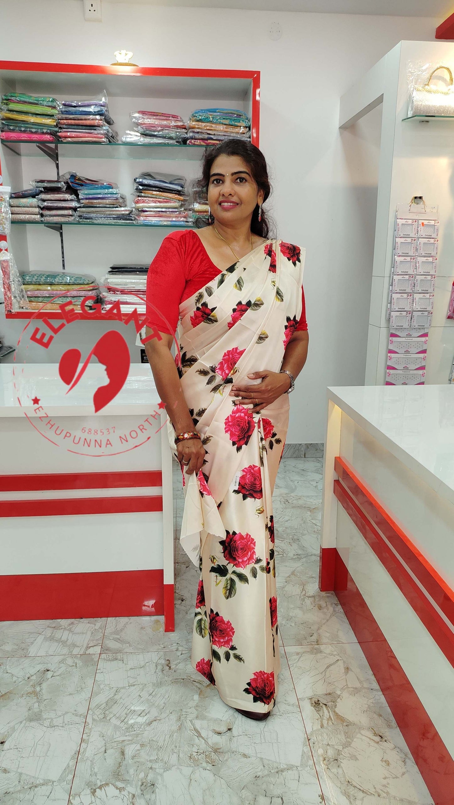 Woman wearing a white Satin Silk Sarees with red floral prints standing in a clothing store with folded sarees on shelves in the background
