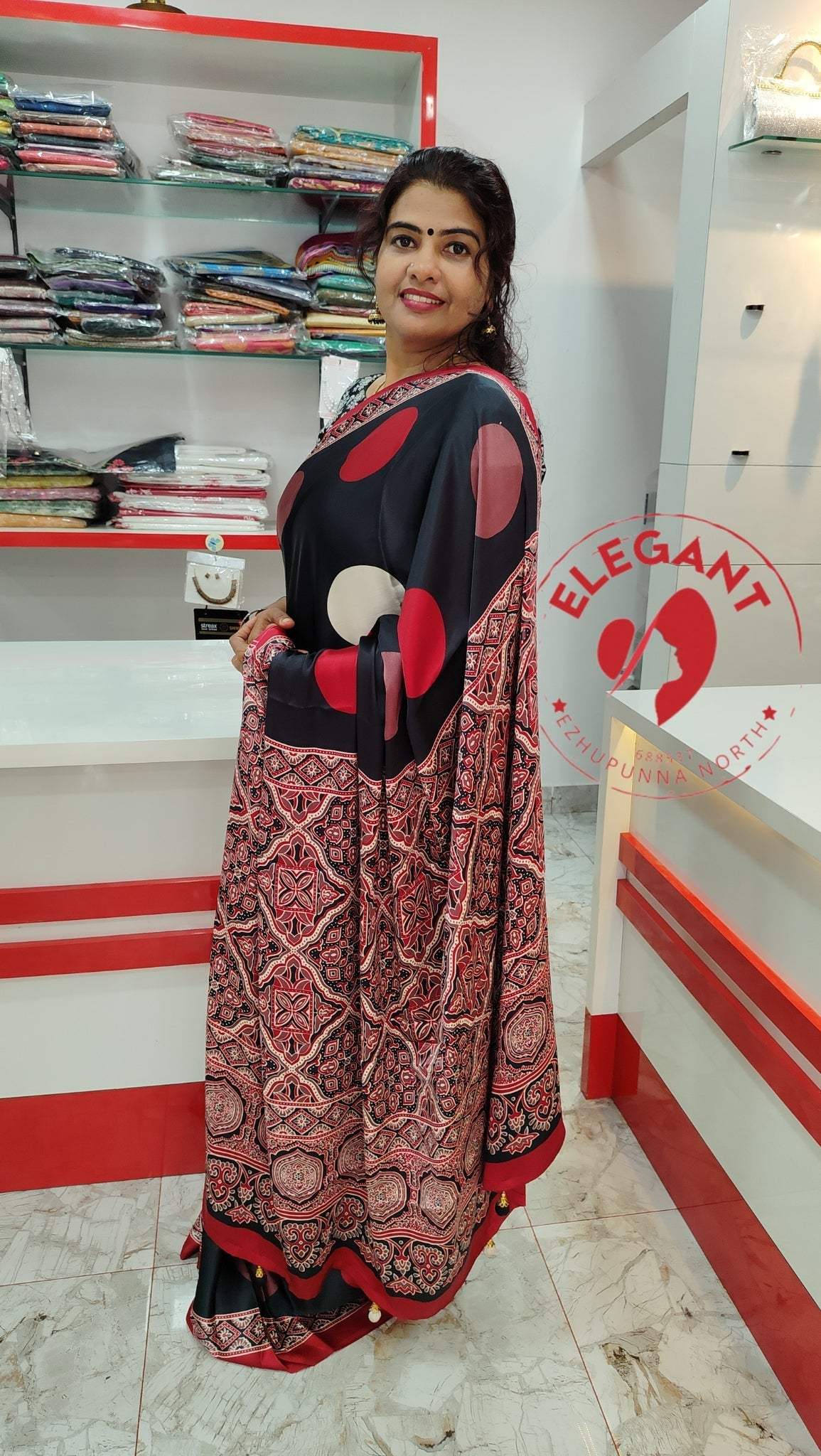 Woman wearing a black and red semi modal silk saree with intricate patterns, standing in a store showroom with shelves of folded sarees in the background.