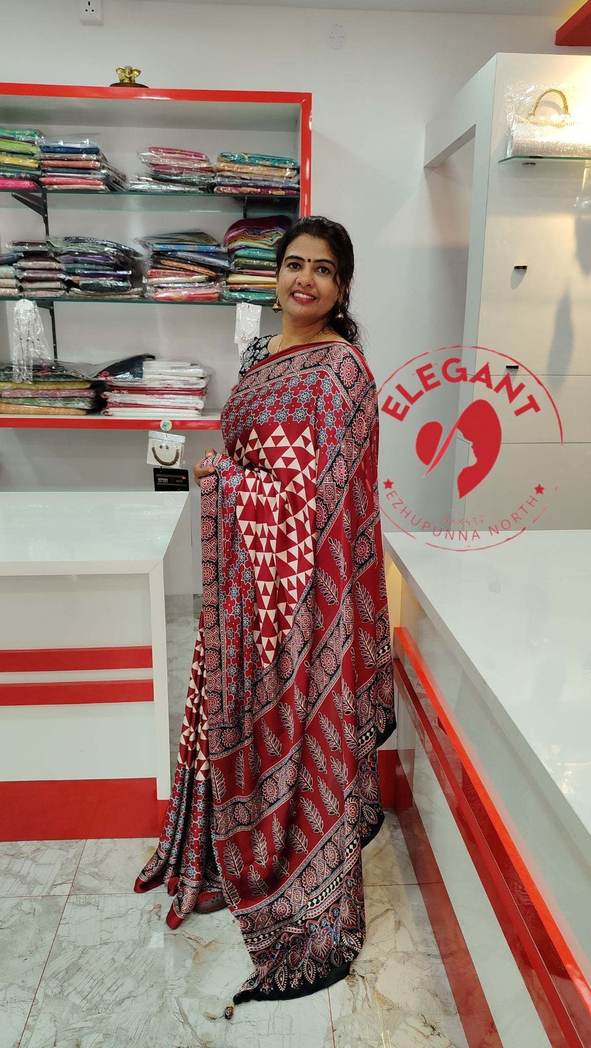 Woman wearing a red and black semi modal silk saree with geometric and floral patterns, standing in a clothing store.