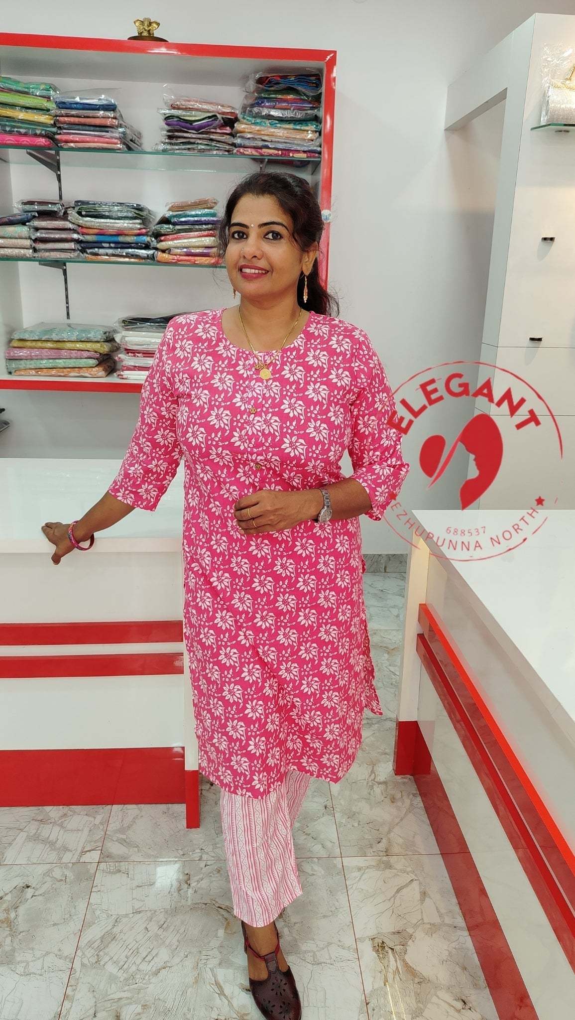 Woman wearing a pink and white Kurthi Set standing in a clothing store with folded garments in the background