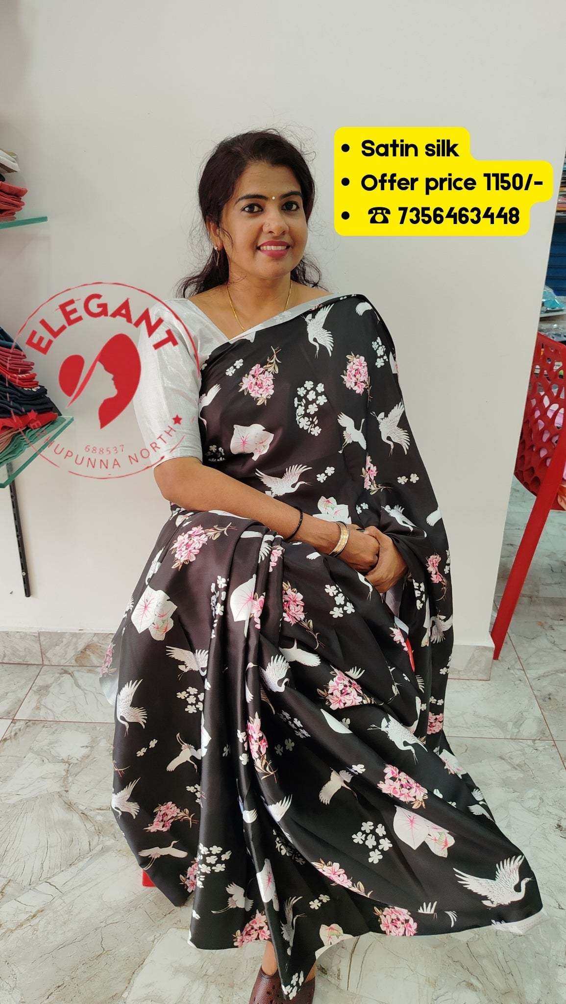 Woman wearing a black satin silk saree with pink and white floral patterns sitting indoors, showcasing satin silk sarees for occasion wear.