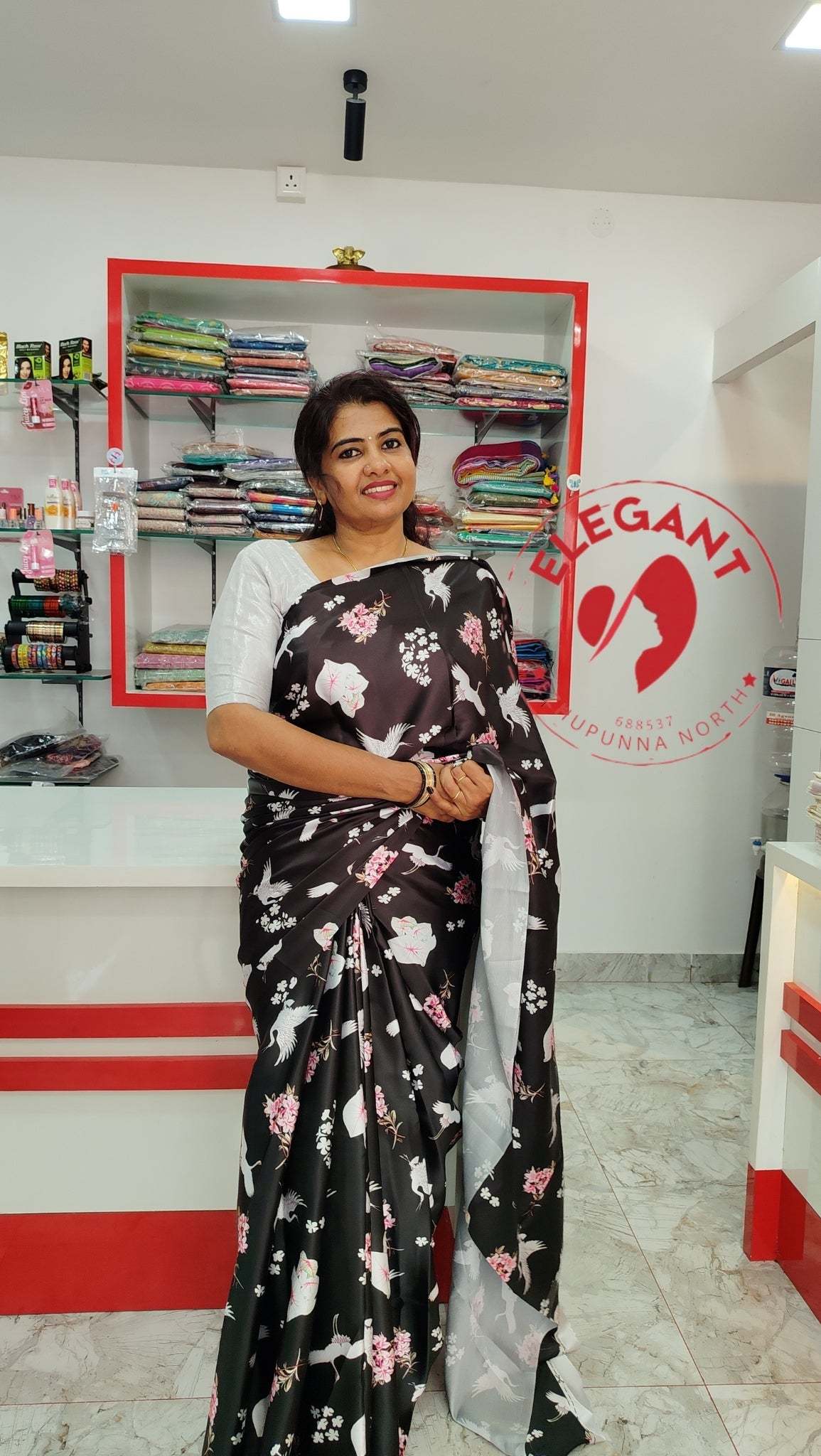 Woman wearing black satin silk saree with white and pink floral patterns in a fabric store displaying Satin Silk Sarees for occasion and party wear