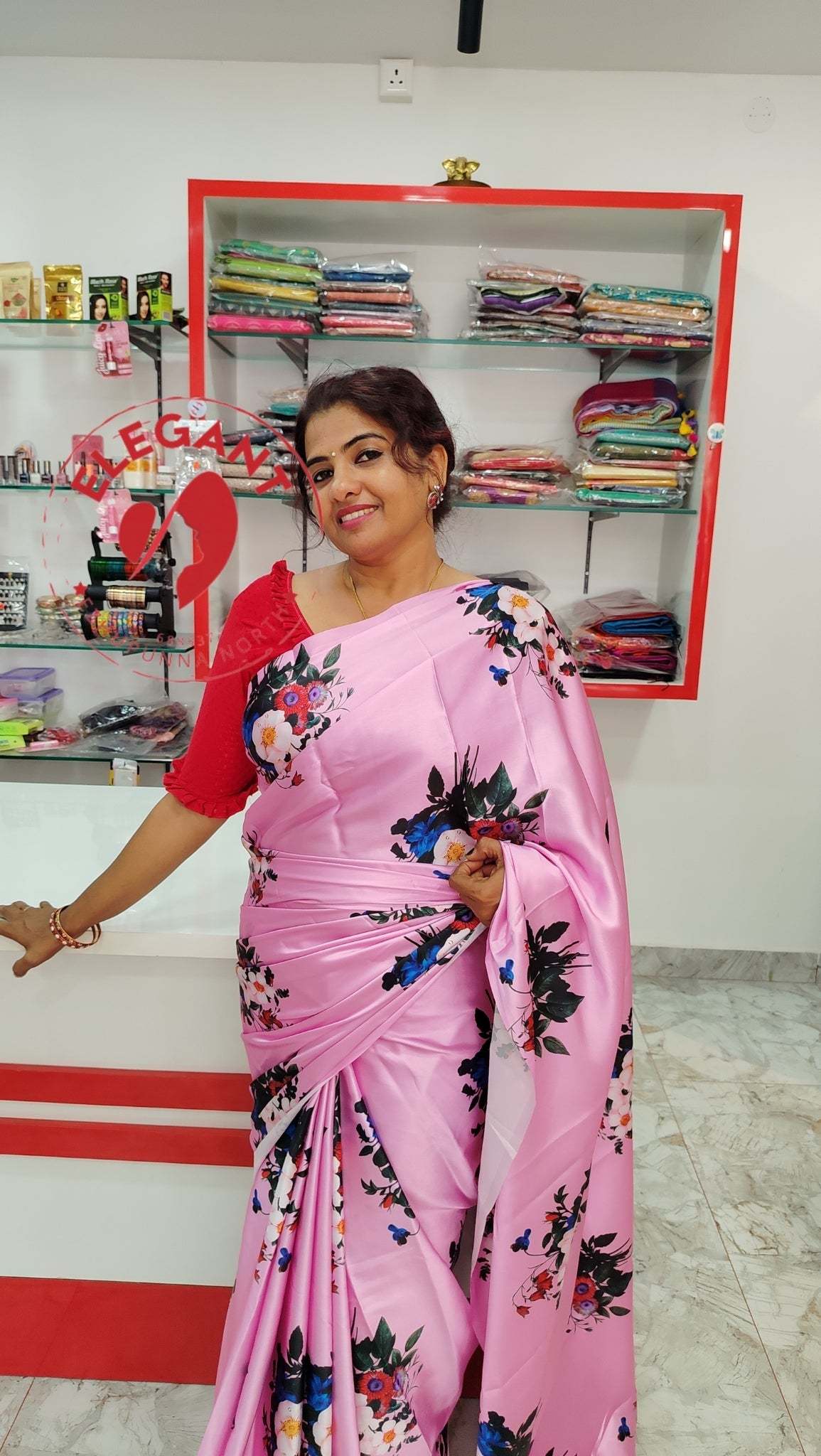 Woman wearing a pink satin silk saree with floral prints standing in a fabric store showcasing satin silk sarees.