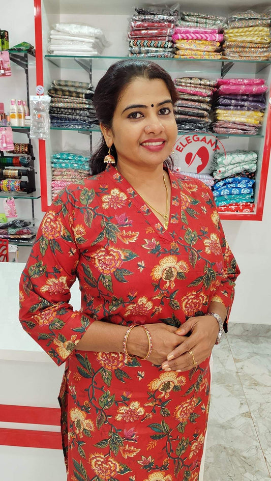Woman wearing red floral Kurthi Set standing in a fabric store with stacked colorful textiles in the background
