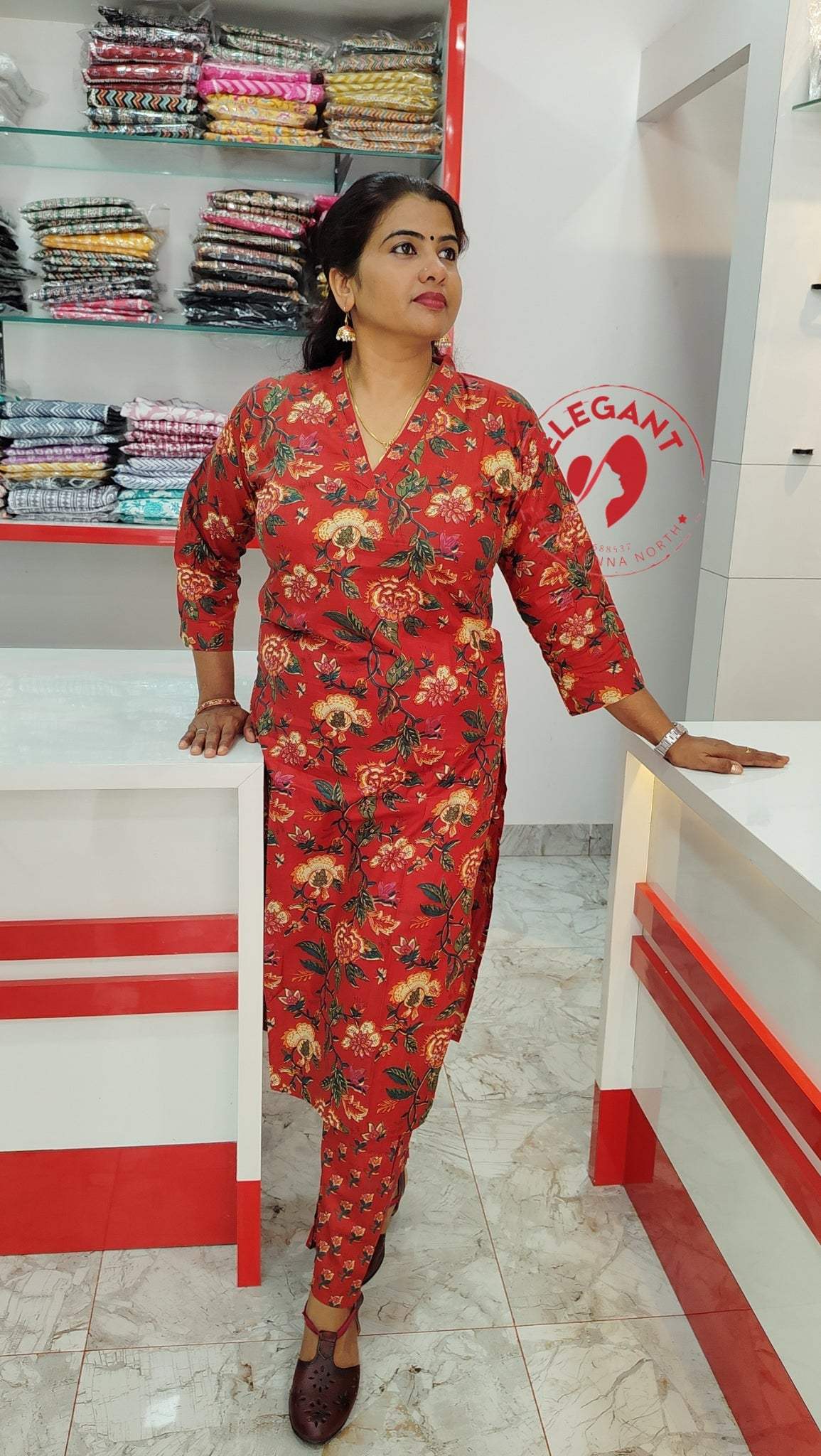 Woman wearing a red floral Kurthi Set standing in a clothing store with folded fabric stacks in the background