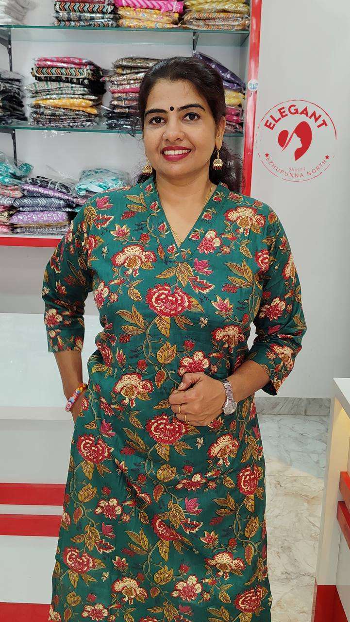 Woman wearing a green floral kurthi set made of lightweight camric cotton in a fabric store with colorful folded fabrics on shelves behind her