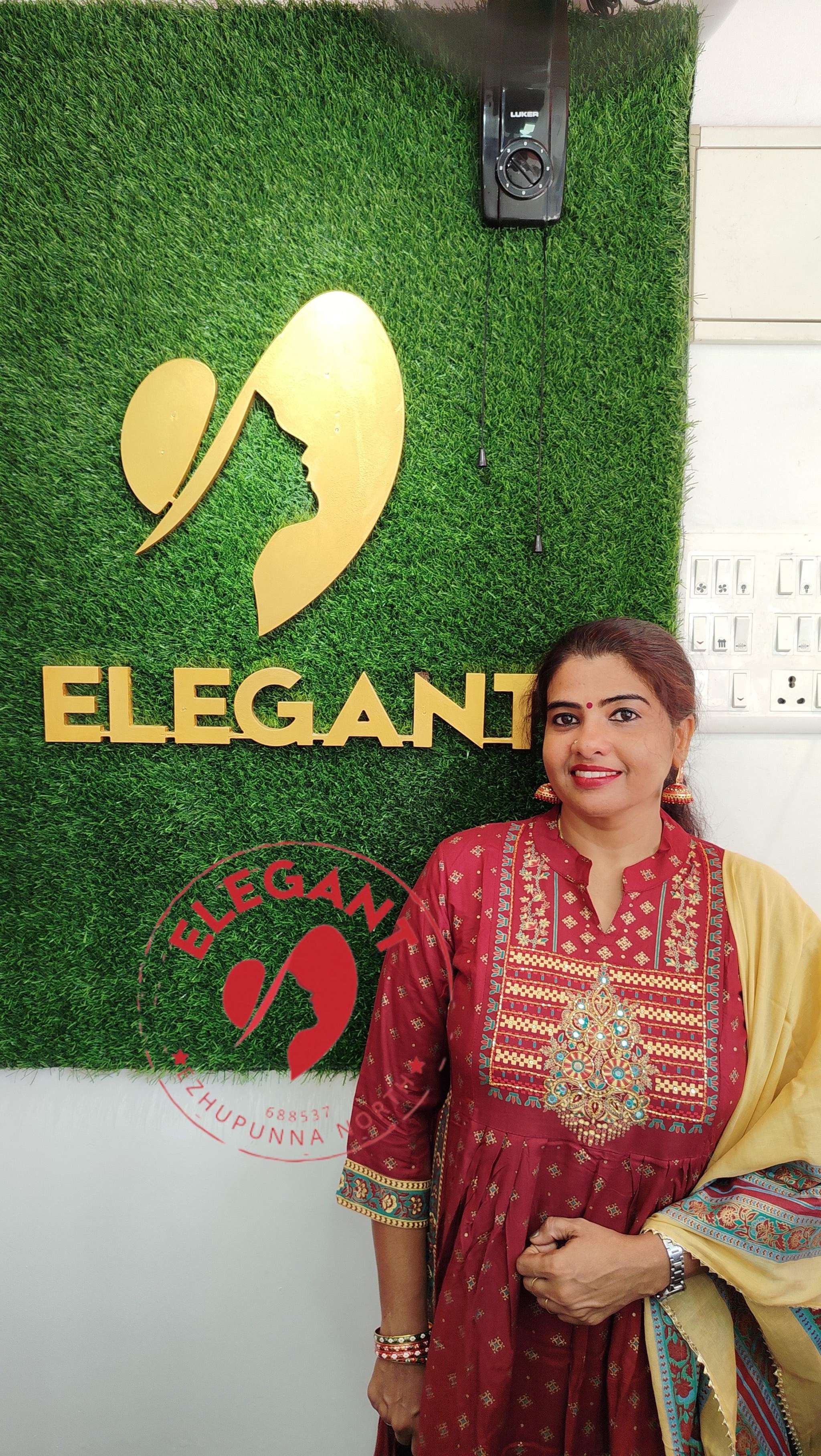 Woman wearing maroon and yellow cotton churidar set standing in front of Elegant store sign on green grass wall