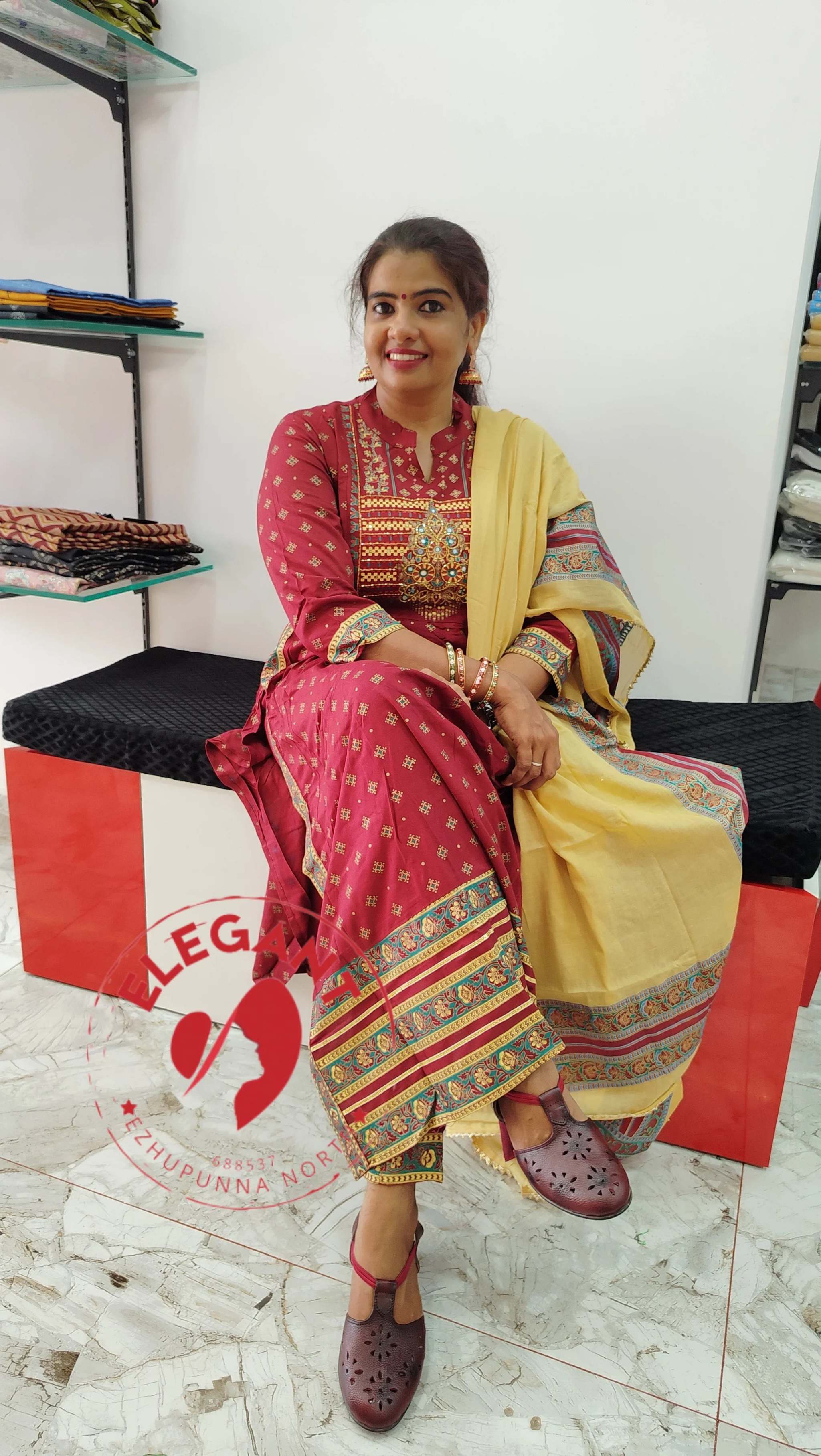 Woman wearing a red cotton churidar set with gold and multicolor patterns, paired with a yellow dupatta, seated indoors.