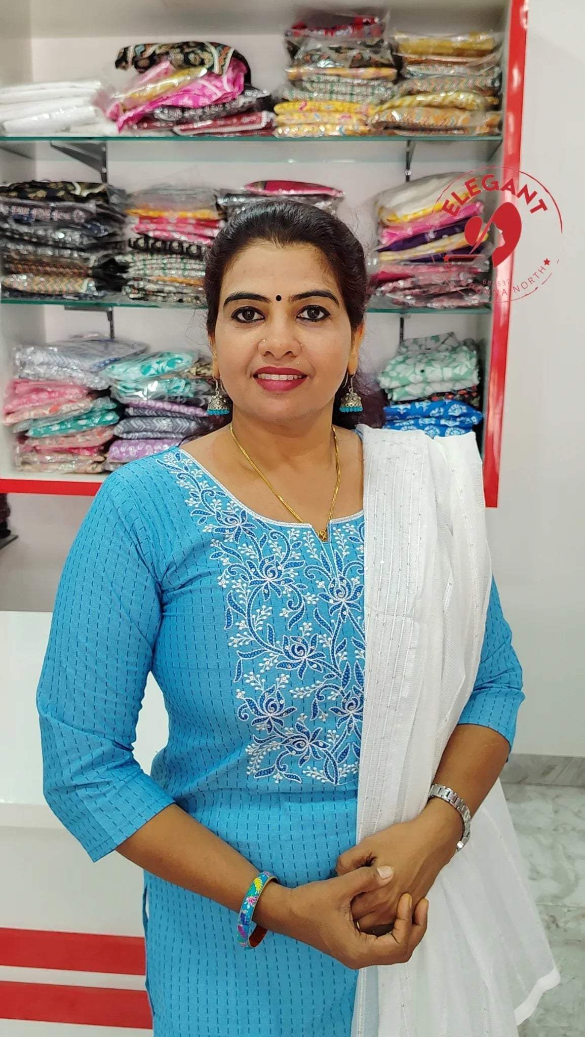 Woman wearing blue silk churidar set with white dupatta standing in front of shelves with folded colorful fabric packs