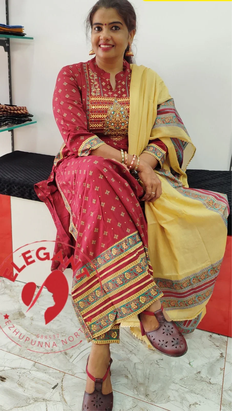 Woman wearing a red cotton churidar set with intricate gold and blue patterns, paired with a yellow dupatta, seated indoors