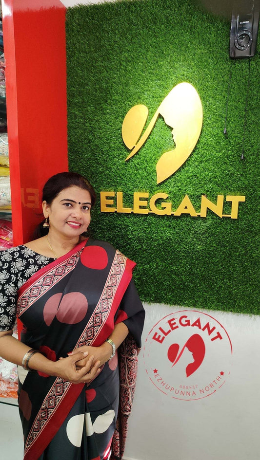 Woman wearing a black and red semi modal silk saree standing in front of Elegant store signage at a retail location.
