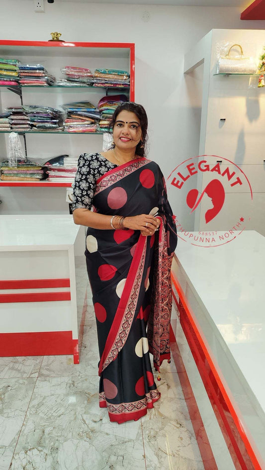 Woman wearing a black semi modal silk saree with red and white polka dots and floral border standing in a saree showroom with shelves of folded fabrics behind her