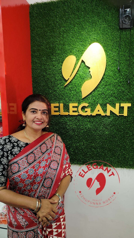 Woman wearing a red and black patterned semi modal silk saree standing in front of Elegant store logo and green wall backdrop