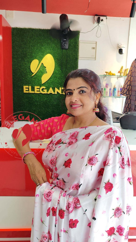 Woman wearing a white satin silk saree with red floral prints standing near a red and green Elegant store sign.