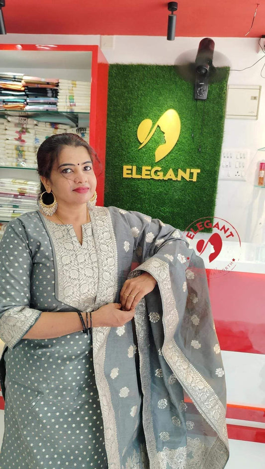 Woman wearing grey silk party wear Churidar with intricate embroidery standing in a store with Elegant brand signage.