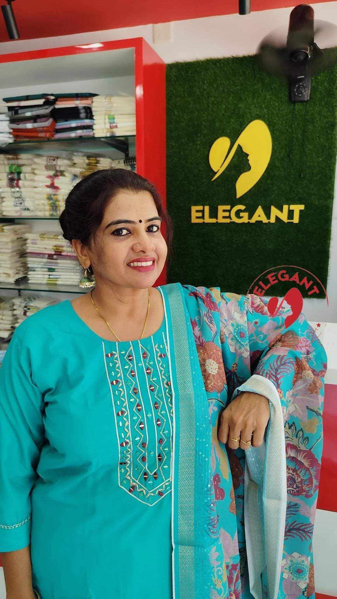 Woman wearing a turquoise Roman Silk Churidar with intricate embroidery and floral patterned dupatta in a clothing store.